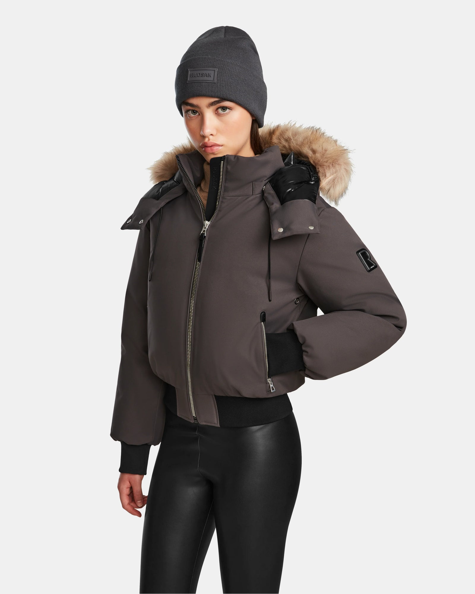 A person in a Rudsak MAUDE - L124522 Grey winter jacket with a fur-trimmed hood, gray beanie, and black faux leather pants stands against a plain white background.