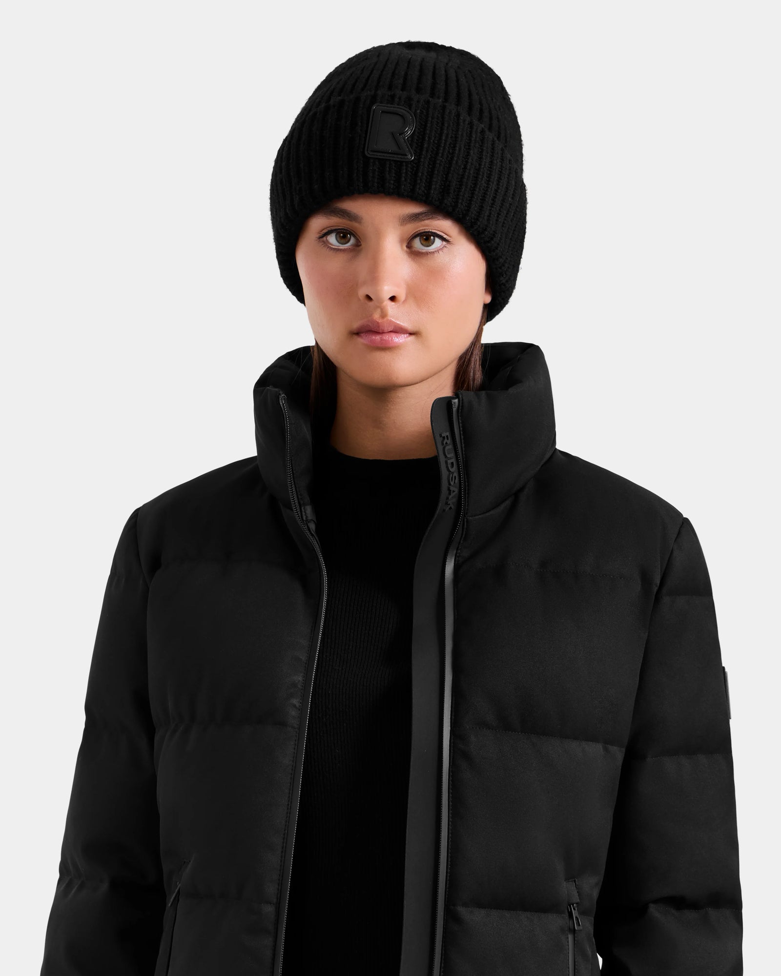A person is wearing the Rudsak MIRIAM MW - L125604 Black women’s down puffer jacket with a high collar and a black knit beanie, facing forward against a plain light background.