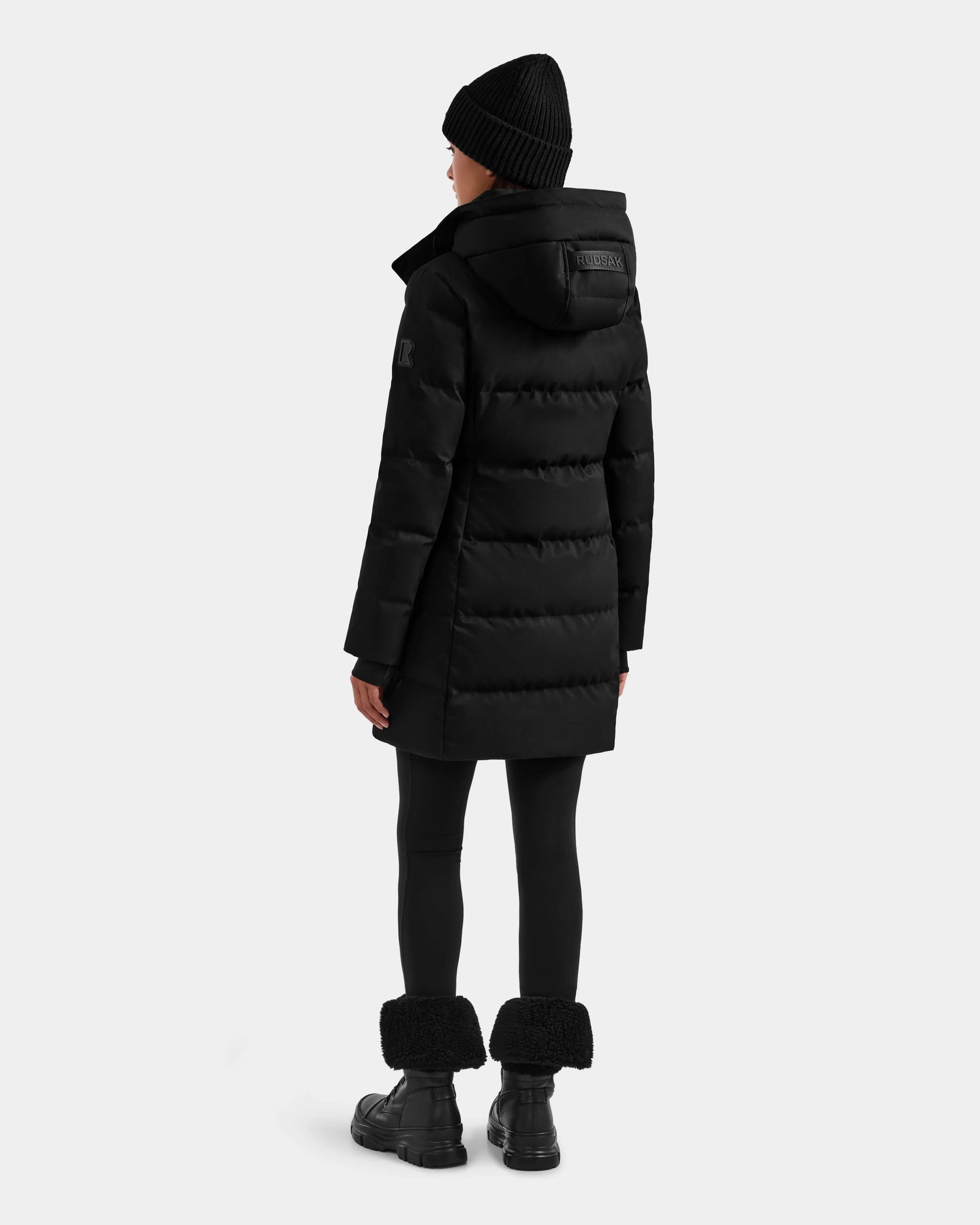 A person wearing Rudsak’s MIRIAM MW - L125604 Black RDS-certified down puffer coat with a hood, a black knit hat, black leggings, and fur-trimmed black boots stands facing away against a plain light gray background.