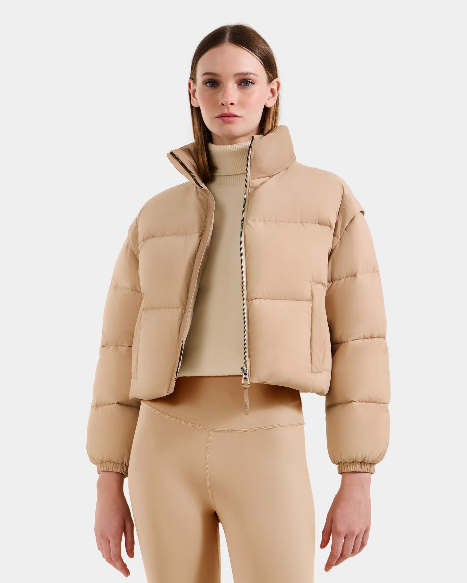 A woman faces forward against a plain white background, wearing the Rudsak OPRAH - L125546 Vanilla cropped down puffer with removable sleeves, paired with a matching turtleneck crop top and high-waisted leggings.