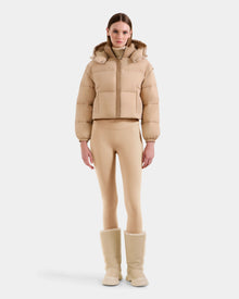 A woman faces forward against a plain white background, wearing the Rudsak OPRAH - L125546 Vanilla beige cropped down puffer, paired with matching beige leggings and fur-lined boots.