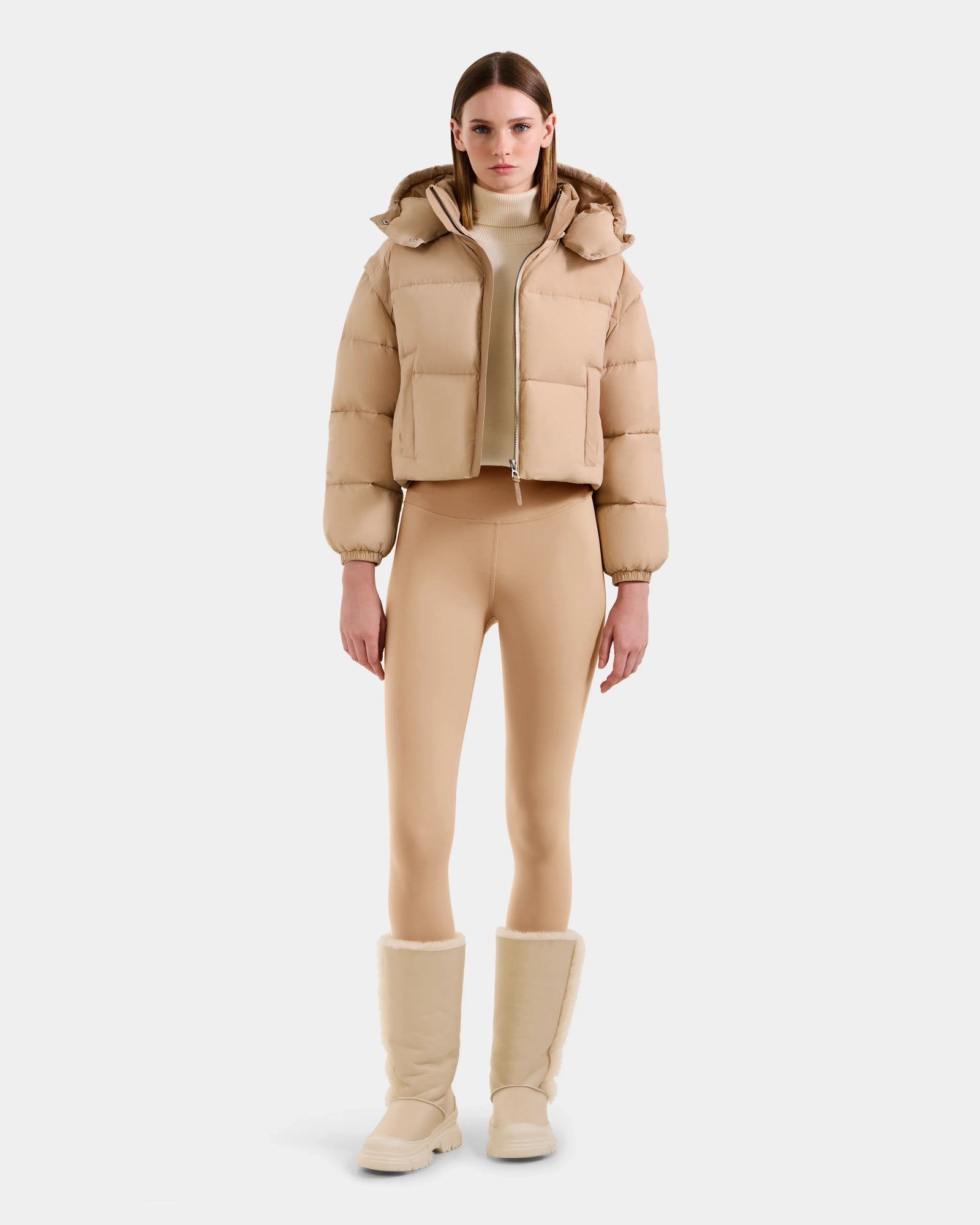 A woman faces forward against a white background, wearing the Rudsak OPRAH - L125546 Vanilla cropped down puffer with removable sleeves, styled with tan fitted leggings, a light turtleneck, and beige fur-lined boots.