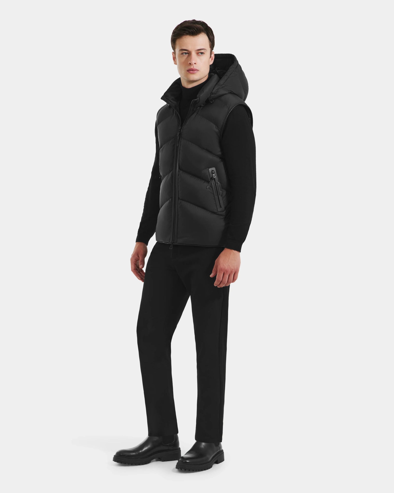 A man faces slightly left, wearing the Rudsak RICHARDSON - M125527 Black men’s hooded vest over a black turtleneck, with black pants and boots, set against a plain white background.