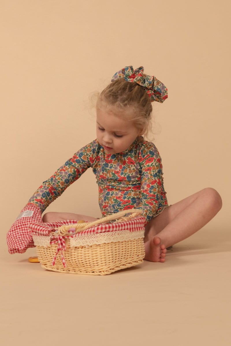 Toddler wearing long-sleeve floral rashguard in red, blue, and gold tones with matching hair accessory.