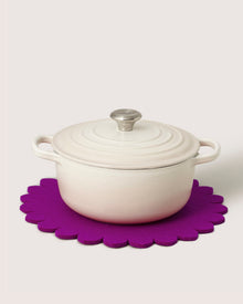 A white ceramic casserole dish with lid rests on a bright purple Merino wool trivet by graf lantz, inspired by Japanese floral crests, providing stylish heat protection against a plain light background.