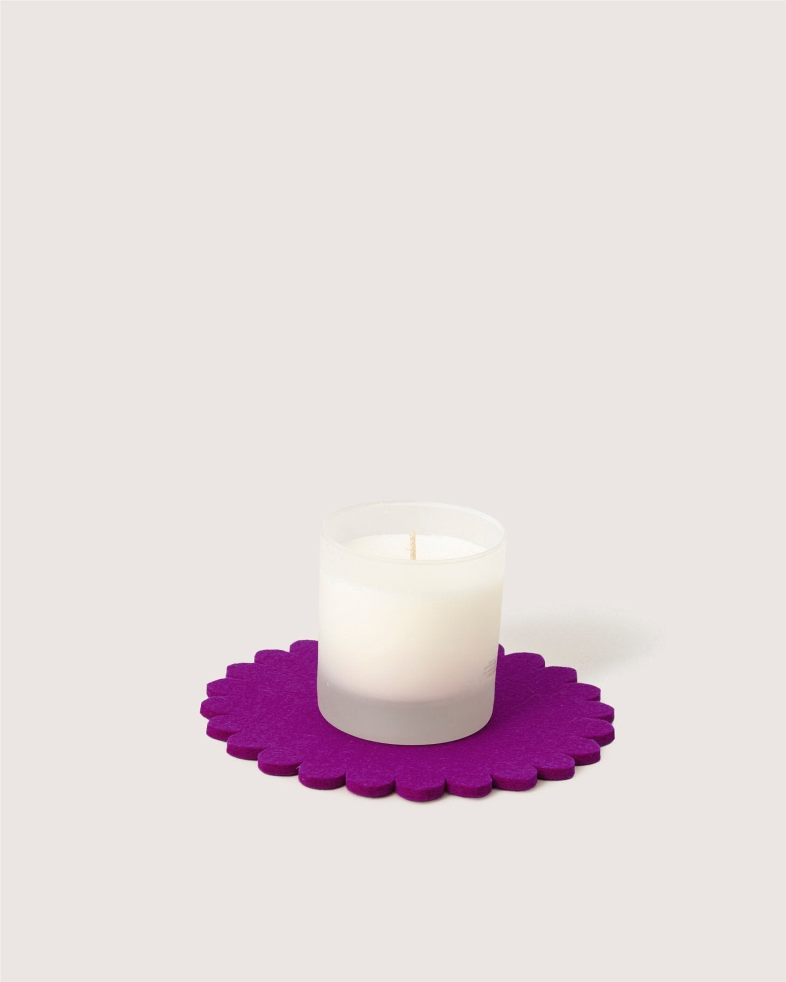 A white frosted glass candle rests on a round, scalloped-edge purple Merino wool trivet by graf lantz, inspired by a Japanese floral crest, set against a plain light background.