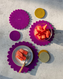 A plate with watermelon and a fork sits on a purple Japanese floral crest placemat; nearby, a glass bowl of watermelon rests on a graf lantz Merino wool trivet, with yellow circular graf lantz coasters on a light gray surface.