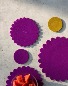 Four graf lantz felt coasters—three purple with scalloped edges and one yellow with a smooth edge—are arranged on a light surface to protect your kitchen. A glass bowl of cubed watermelon is at the bottom left of the image.