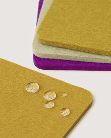 Four square Graf Lantz Merino wool coasters in mustard yellow, off-white, and purple are stacked with one yellow coaster in front. Water drops on the felt surface showcase their water resistance.
