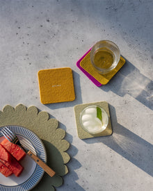 A plate with watermelon slices and a fork sits next to two drinks on vibrant Graf Lantz Merino wool coasters atop a textured gray surface with long shadows.