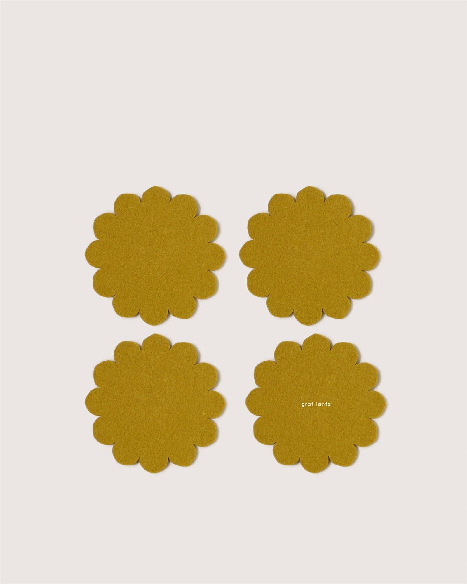 Four mustard yellow, scalloped-edge Merino wool coasters by Graf Lantz, inspired by Japanese floral crests, are arranged in a grid on a white background. One coaster features subtle graf lantz text, adding stylish surface protection.