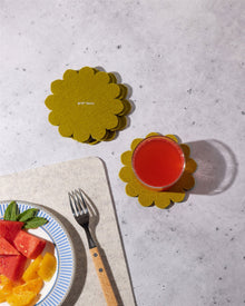 A glass of red juice sits on a yellow flower-shaped Merino wool coaster by Graf Lantz, inspired by Japanese floral crests. Nearby, more coasters are stacked for surface protection, with a plate of fruit and fork partially visible.
