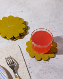 A glass of pink-red drink rests on a yellow Graf Lantz Japanese floral crests coaster, with more Graf Lantz Merino wool coasters stacked nearby, next to a fork on a napkin and a blue-striped plate atop a light-colored surface.