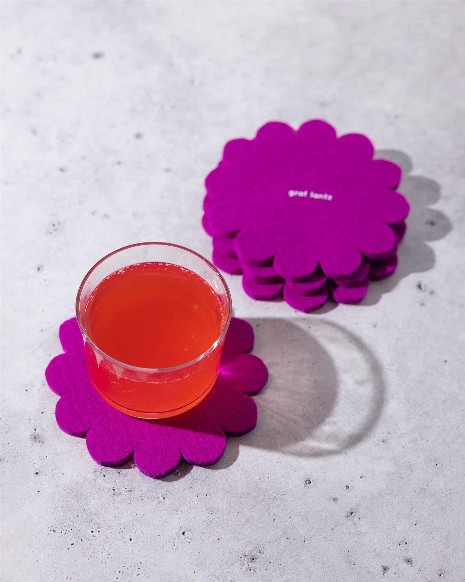A glass of red-orange drink rests on a bright purple Japanese floral Merino wool coaster by Graf Lantz, protecting the surface, with a stack of matching flower-shaped coasters nearby on a light gray background.