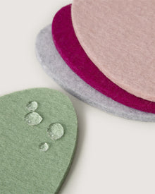Close-up of stacked Graf Lantz round felt coasters in pastel shades—green, gray, pink, magenta. The green one glistens with water droplets. Made of Merino wool for a soft, absorbent look similar to egg coasters.
