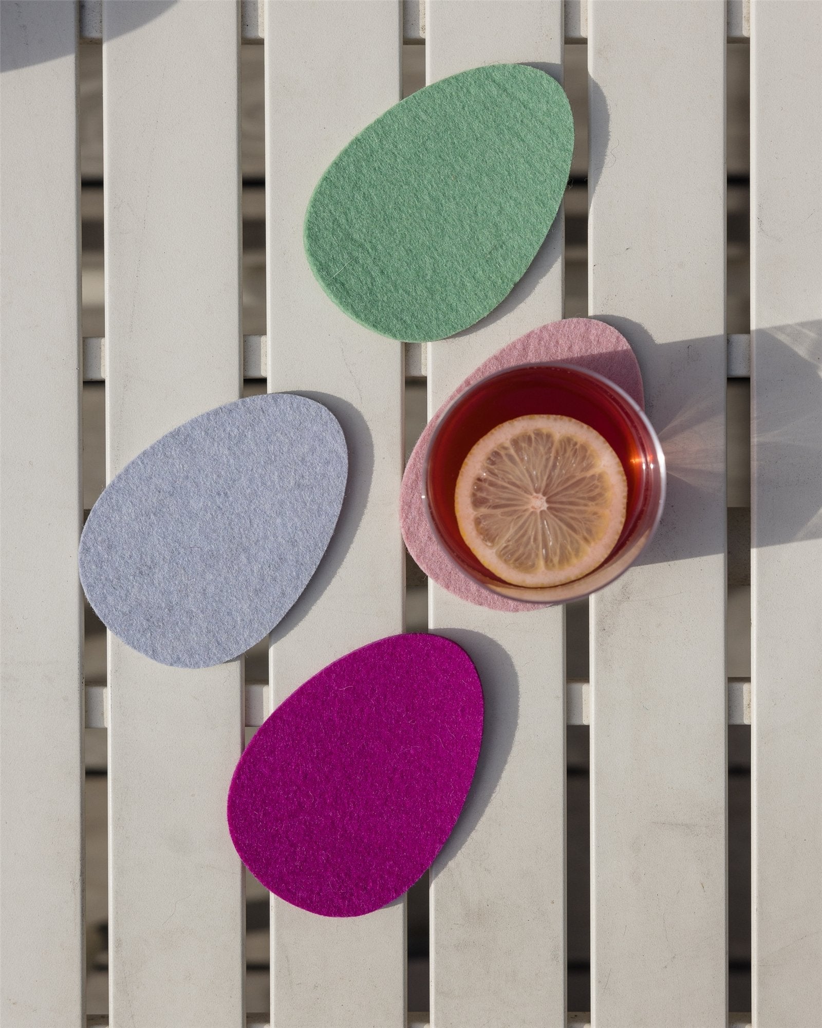 A glass of tea with a lemon slice rests on a pink felt coaster on a white wooden surface. Nearby, three pastel egg-shaped coasters in green, blue, and magenta add charm, crafted from soft Merino wool by Graf Lantz.