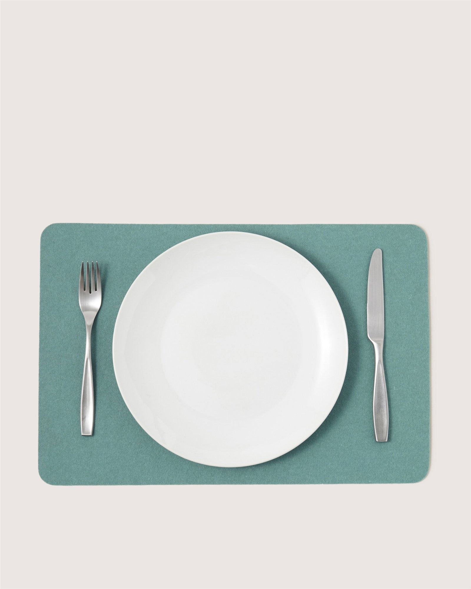A white empty plate sits centered on a green Graf Lantz Merino wool placemat, with a silver fork to the left and knife to the right, creating stylish designer table decor against a light background.
