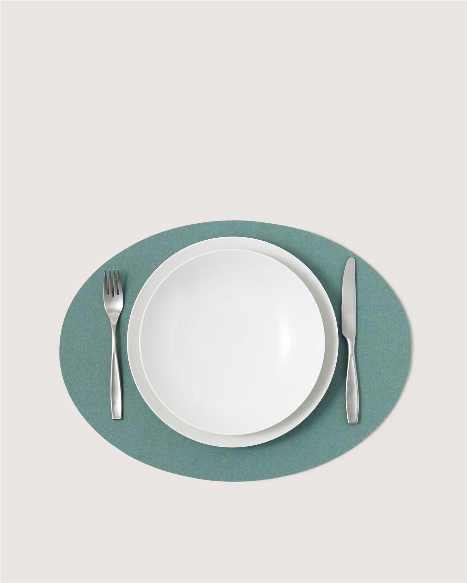A white plate and bowl sit on a green Graf Lantz Merino wool placemat, flanked by a silver fork and knife, all arranged neatly against a plain background for an elegant designer table setting.