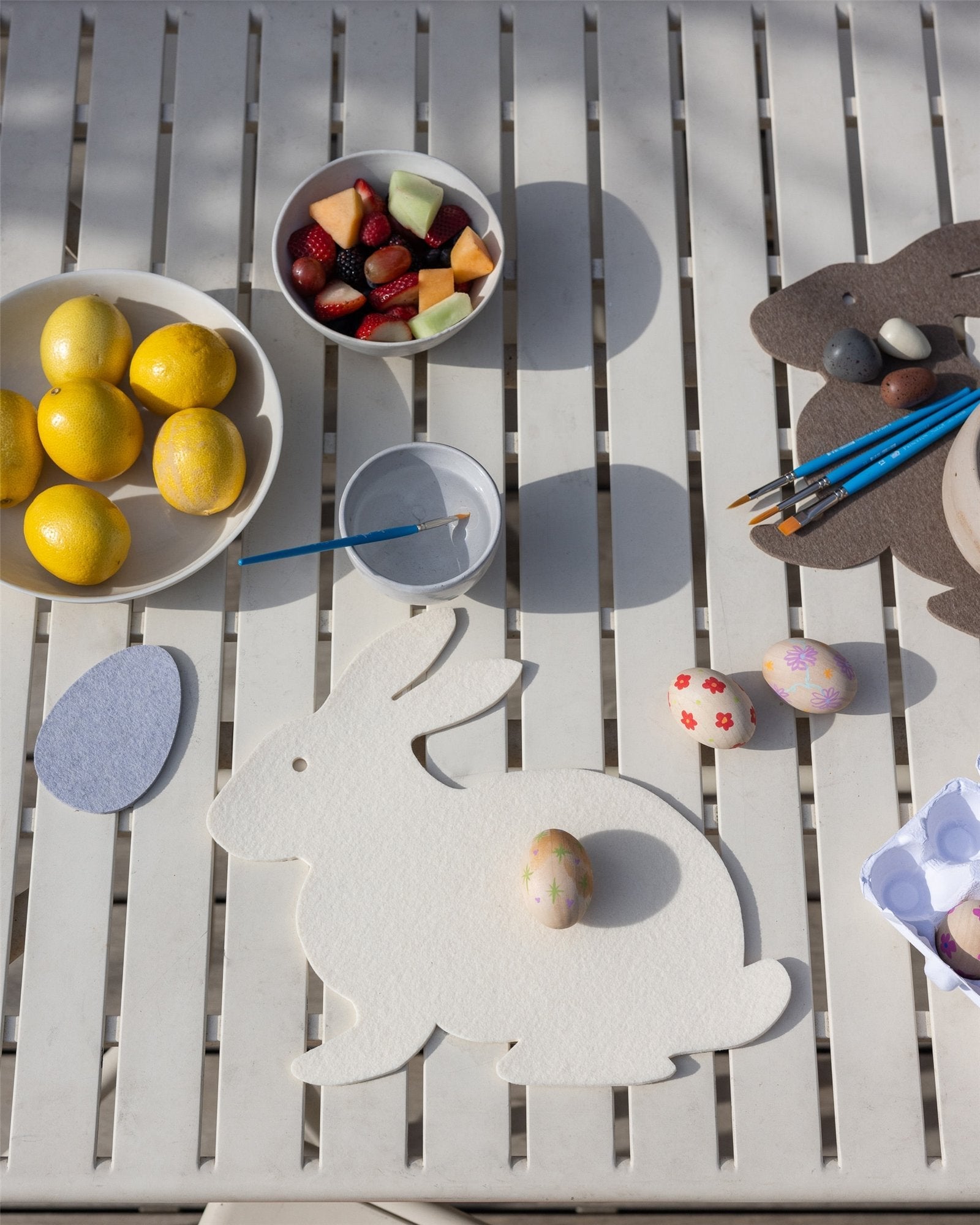 A Graf Lantz Merino Wool Bunny Trivet highlights a table of Easter craft supplies, including decorated eggs, paintbrushes, a palette, and bowls of lemons and mixed fruit, creating a festive and creative atmosphere.