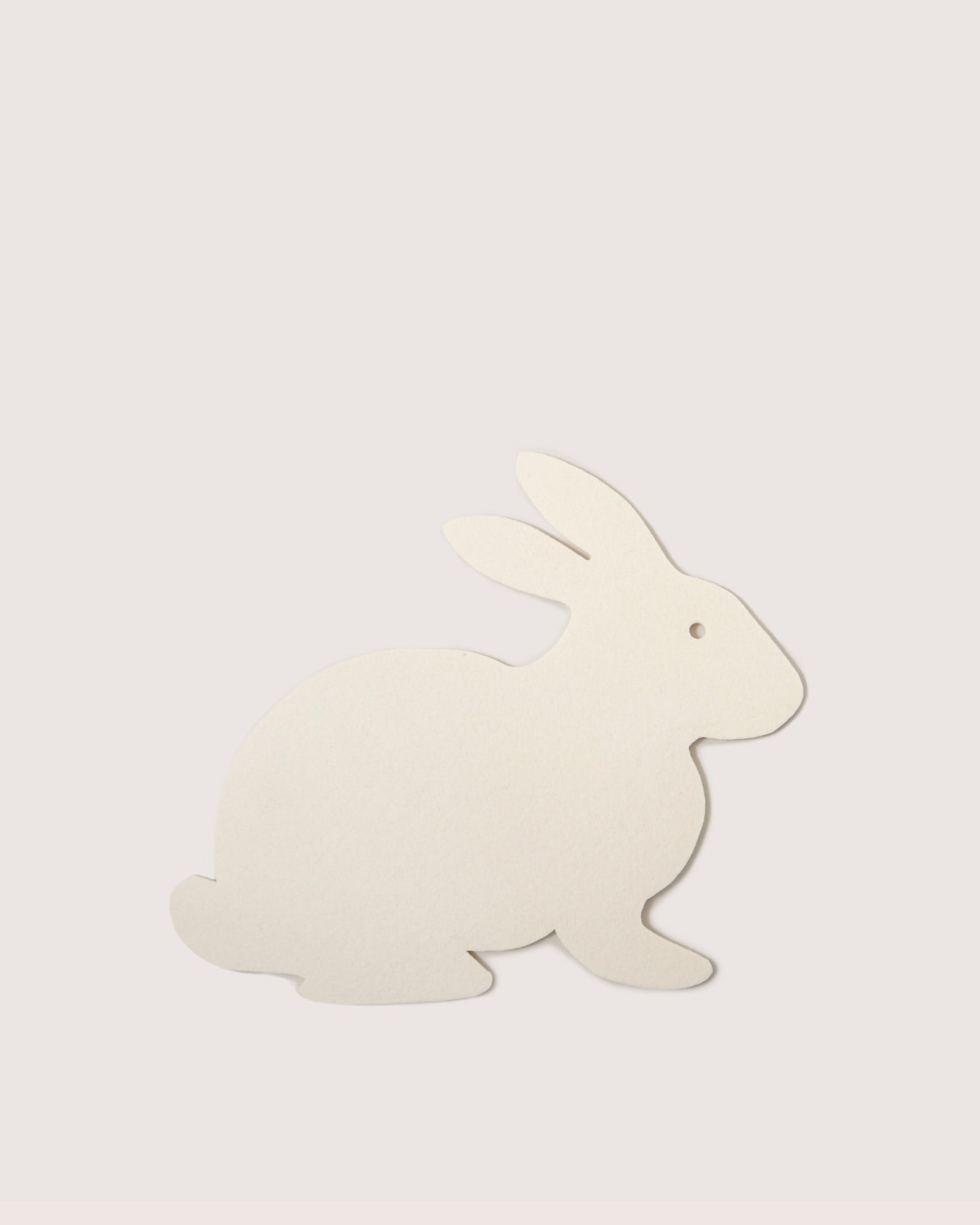 A season-exclusive Graf Lantz trivet features a simple beige flat cut-out rabbit design on a light gray background. Resembling thick paper or thin cardboard, it faces right with minimal detail, similar to Merino wool.