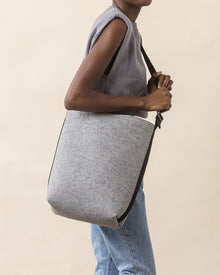 A person holds a large gray felt shoulder bag by graf lantz with a black strap, highlighting everyday functionality, while wearing a sleeveless gray top and light blue jeans against a plain beige background.