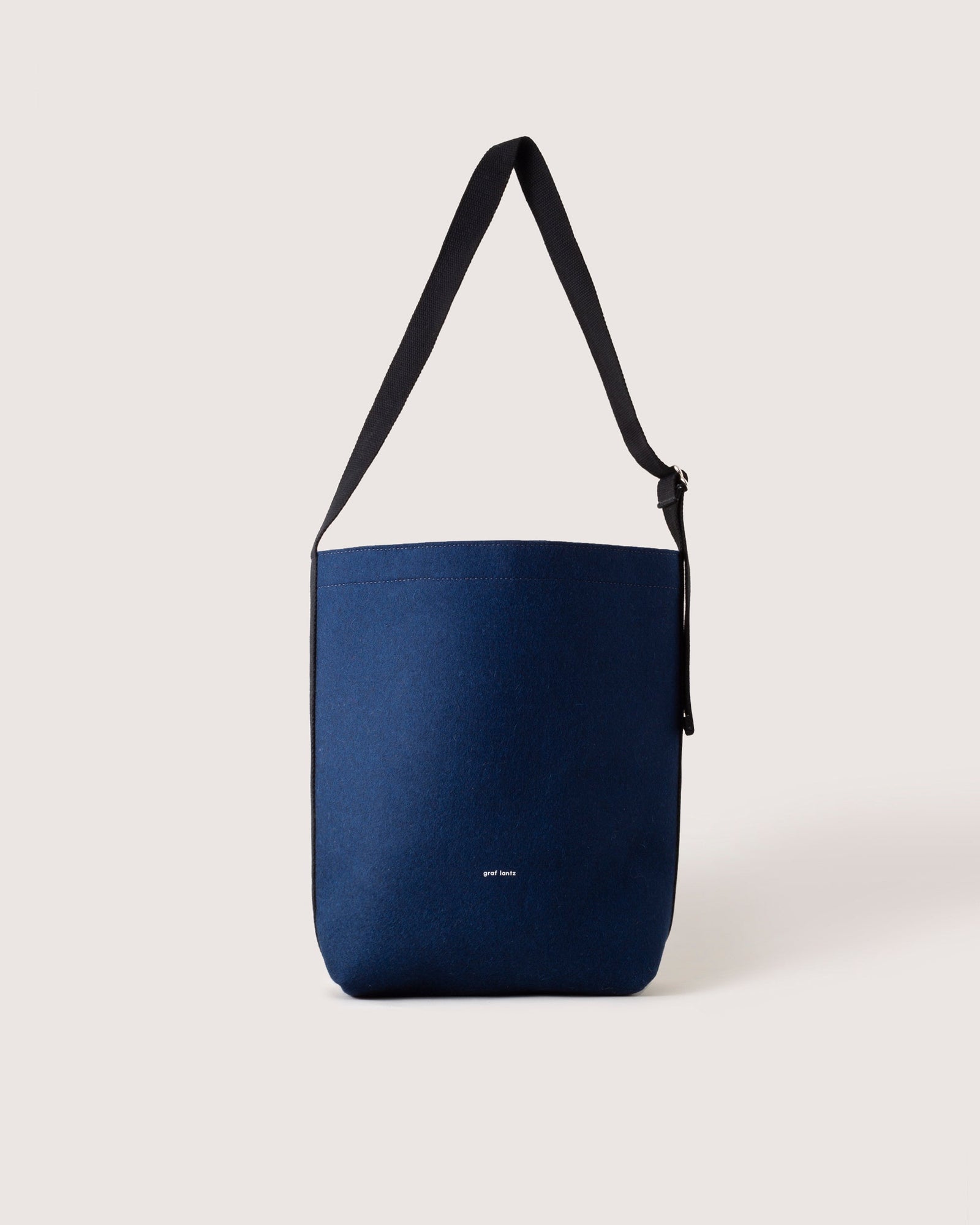 A minimalist navy blue shoulder bag from graf lantz, featuring a black strap and crafted from sustainable materials, stands out against a neutral background with a small white logo near the bottom.