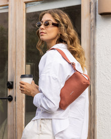 A wavy-haired individual in sunglasses, a white Merino Wool shirt, and cream pants holds a reusable coffee cup. They carry Graf Lantzs rust-colored Campus Shoulder Bag and stand before a glass door.