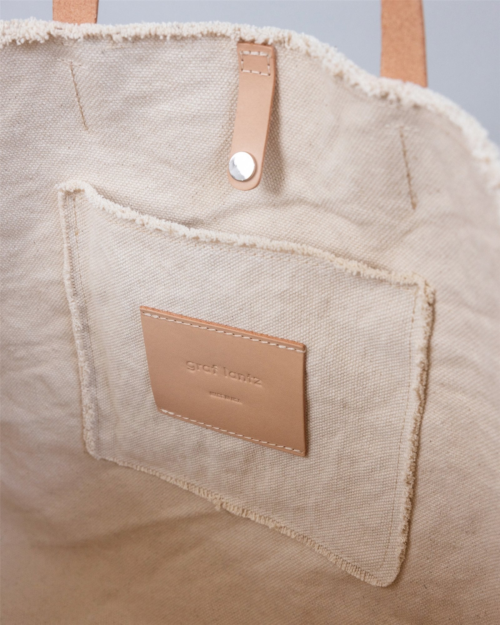 Close-up of the Graf Lantz Hana Canvas Totes beige interior, highlighting a stitched pocket, branded leather patch, leather straps, and a metal snap for timeless style.