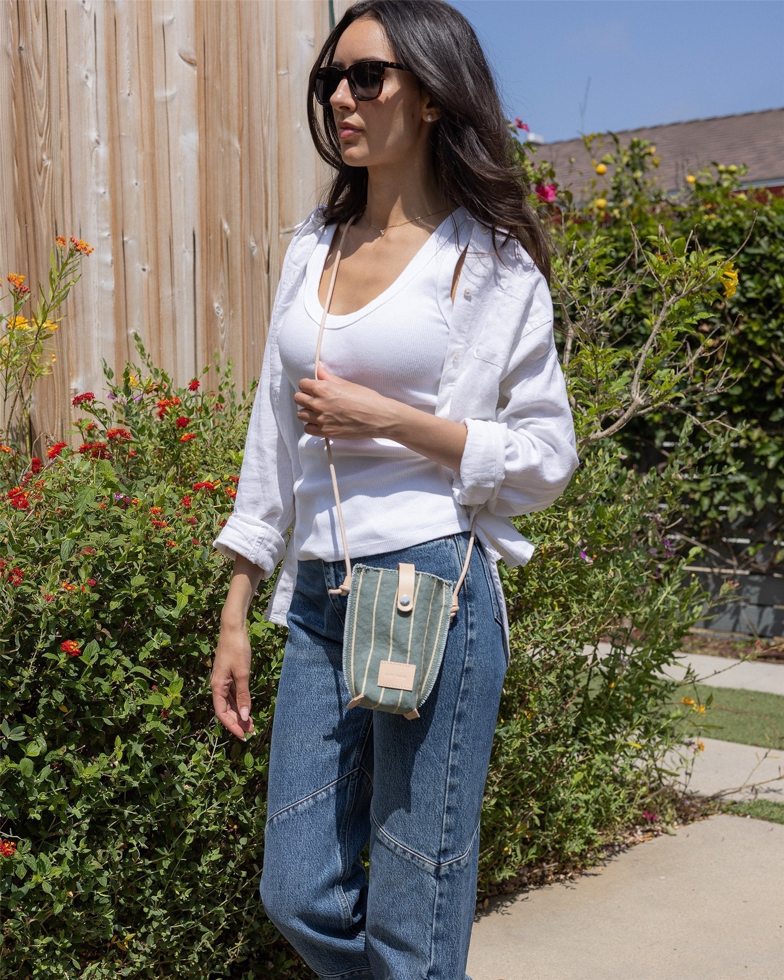 A woman wearing sunglasses, a white shirt over a tank top, and blue jeans strolls outdoors by green bushes and a wooden wall, carrying a compact Graf Lantz crossbody bag for a stylish hands-free look.