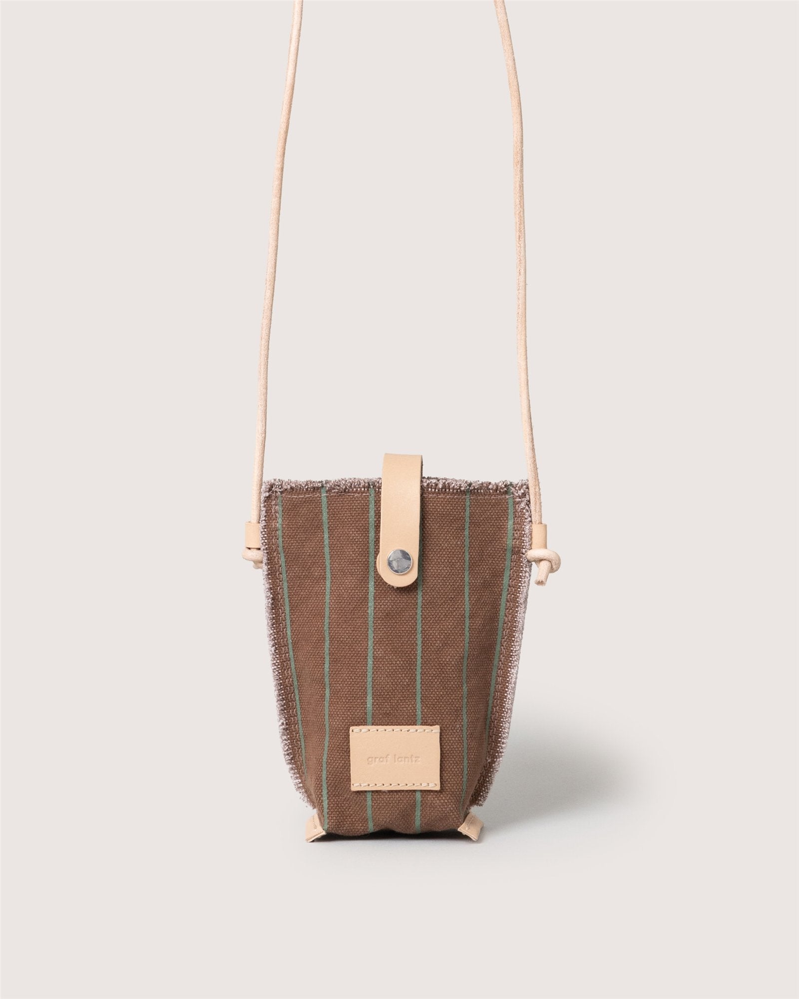 The Graf Lantz Hana Canvas Phone Crossbody is a compact bag in brown fabric with thin green stripes, beige leather strap and trim, flap snap closure, and a rectangular front label. The background is plain light beige.