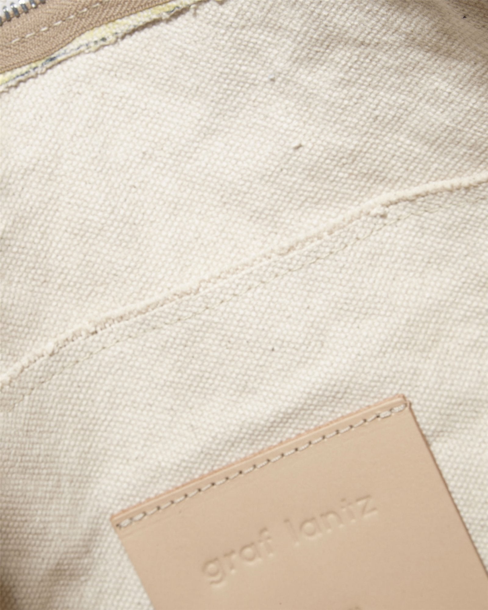 Close-up of beige canvas with a stitched seam and tan Vachetta leather patch embossed with Graf Lantz, sewn onto the fabric. It hints at the timeless elegance of a Hana Hobo bag. The top edge shows part of a zipper.