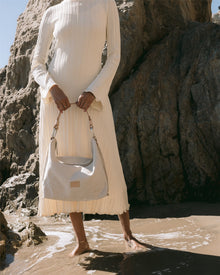 A person stands barefoot on wet sand with waves washing over, holding a Graf Lantz Vachetta leather shoulder bag. They wear a long-sleeved, pleated cream dress. Large rocks are in the background as sunlight casts soft shadows.