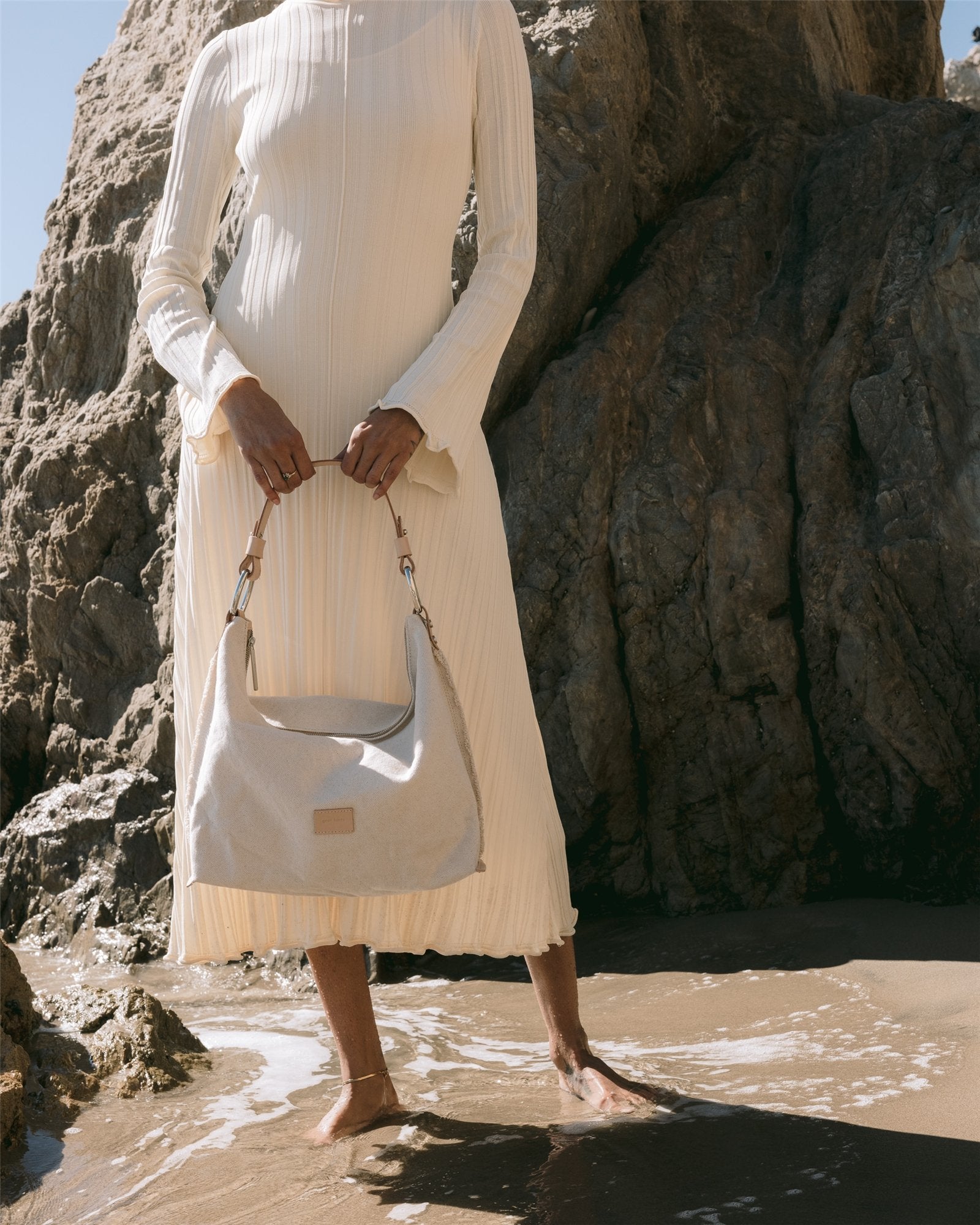 A person stands barefoot on wet sand with waves washing over, holding a Graf Lantz Vachetta leather shoulder bag. They wear a long-sleeved, pleated cream dress. Large rocks are in the background as sunlight casts soft shadows.