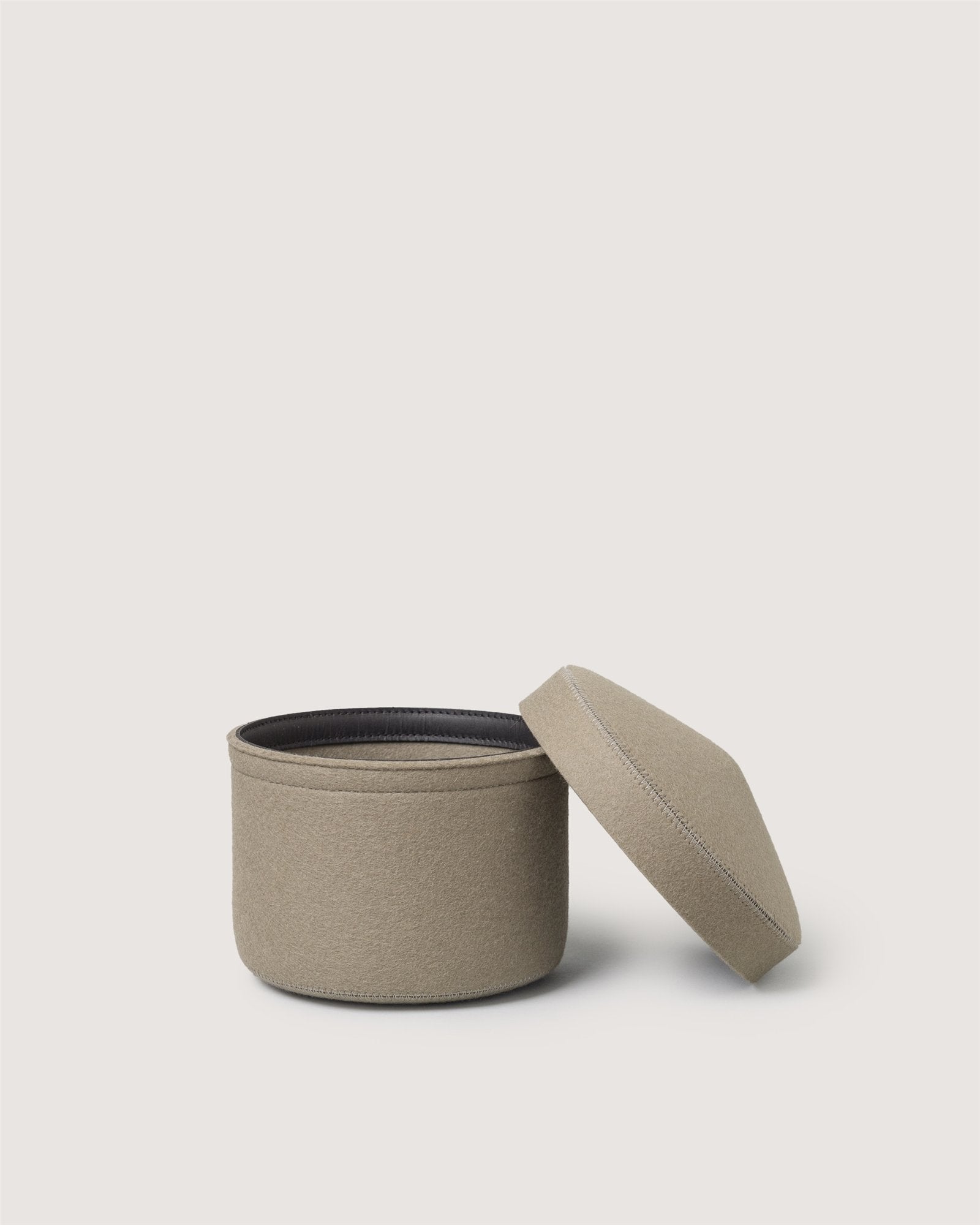 A round, beige Merino wool bin by Graf Lantz with a fitted lid sits slightly askew, revealing its dark interior against a plain light background—a stylish and sustainable storage solution for any space.