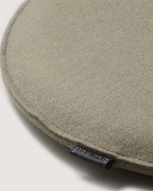 Close-up of a round, light gray Merino wool cushion by Graf Lantz with a small fabric tag on the edge. The background is plain and off-white.