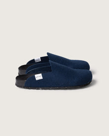 A pair of graf lantz dark blue felt slippers with black recycled rubber soles and a cork footbed, featuring a small white tag on the side, are placed on a light gray background.