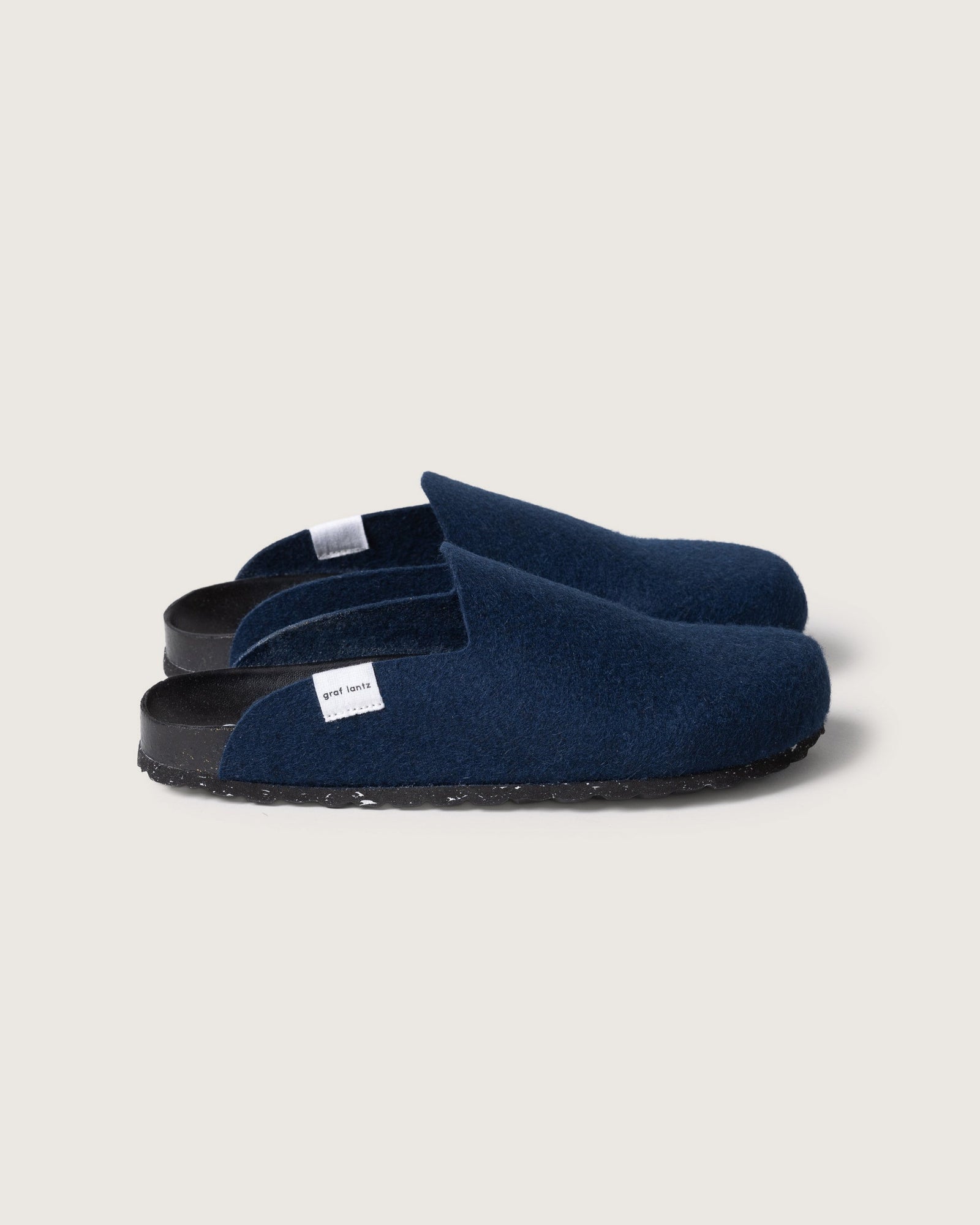 A pair of graf lantz dark blue felt slippers with black recycled rubber soles and a cork footbed, featuring a small white tag on the side, are placed on a light gray background.