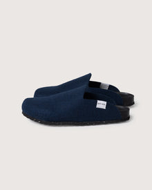 A minimalist pair of dark blue slip-on slippers from graf lantz is shown against a plain background. Crafted from Merino wool, they feature black recycled rubber soles and a small white tag for eco-friendly comfort.