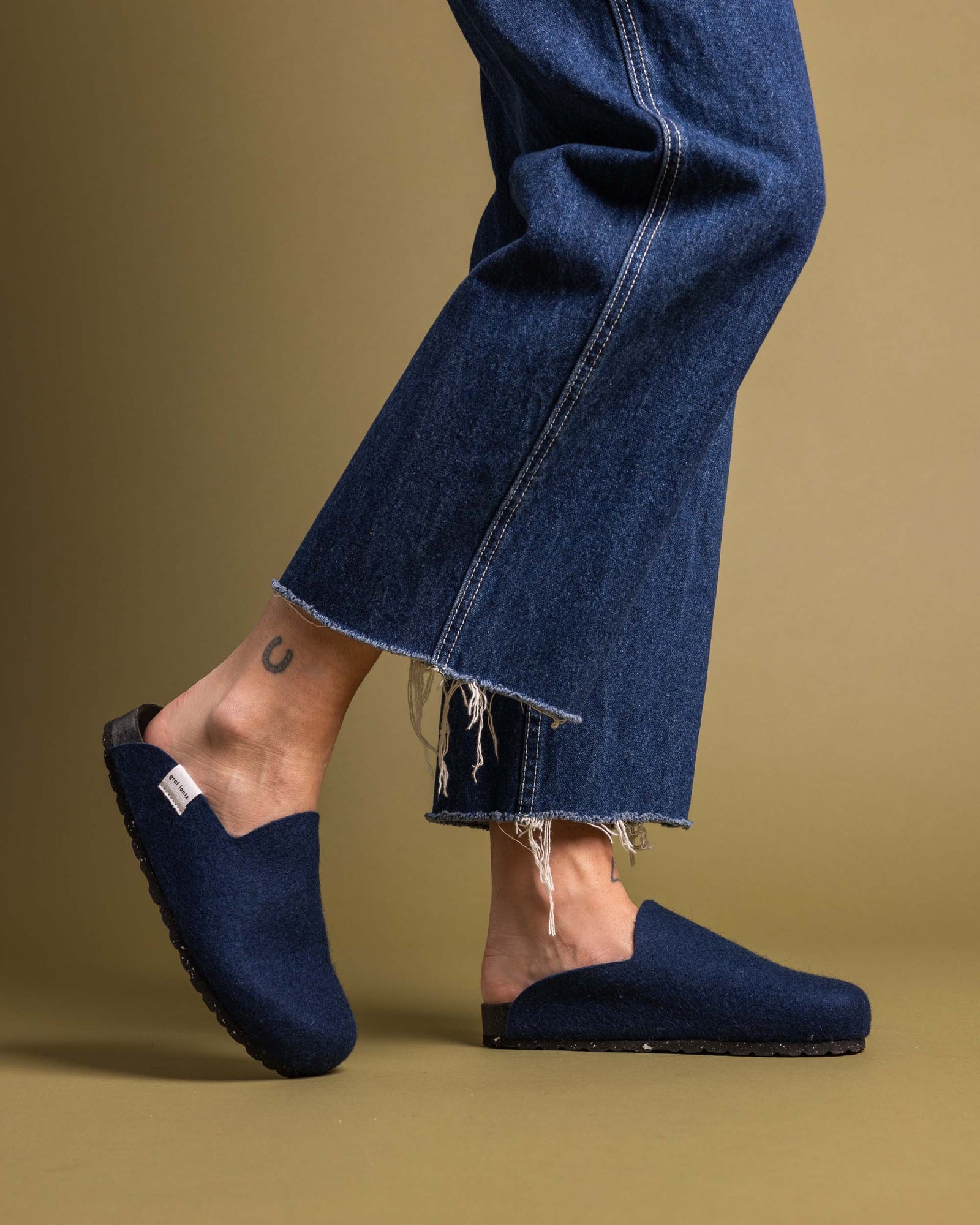 The person showcases graf lantzs dark blue slip-on shoes, featuring a recycled rubber sole, paired with frayed hem jeans against a beige backdrop. A small tattoo peeks out above the ankle.