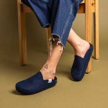 A person sits on a wooden chair wearing graf lantz dark blue felt slippers with recycled rubber soles, frayed hem blue jeans, and a small triangle tattoo on their right ankle. The warm beige background enhances the cozy ambiance.