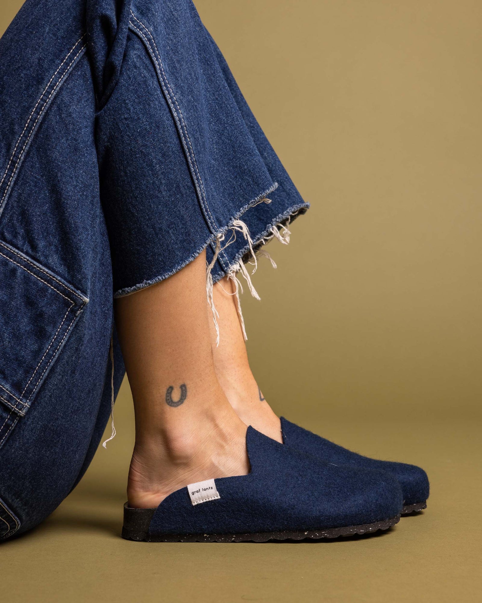 Close-up of a person wearing frayed hem blue jeans and graf lantz blue slip-on shoes with a recycled rubber sole, showcasing a small horseshoe tattoo on one ankle against an olive-tan background.