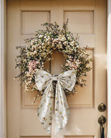 An Easter Basket and Wreath Bow in Easter Frolic with a large white bow featuring a pastel floral pattern is tied at the bottom of a wreath made of small white and pink flowers, hanging on a beige door.