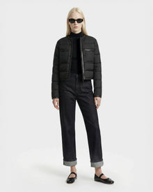 A person with long blonde hair wears black sunglasses and the Rudsak MARKLE - 8125033 Black women’s cropped down jacket, paired with a black turtleneck, dark cuffed jeans, and black flats against a plain white background.