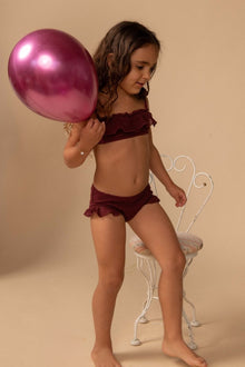 Child wearing maroon crochet bandeau bikini top and ruffled swim shorts with scalloped edges.