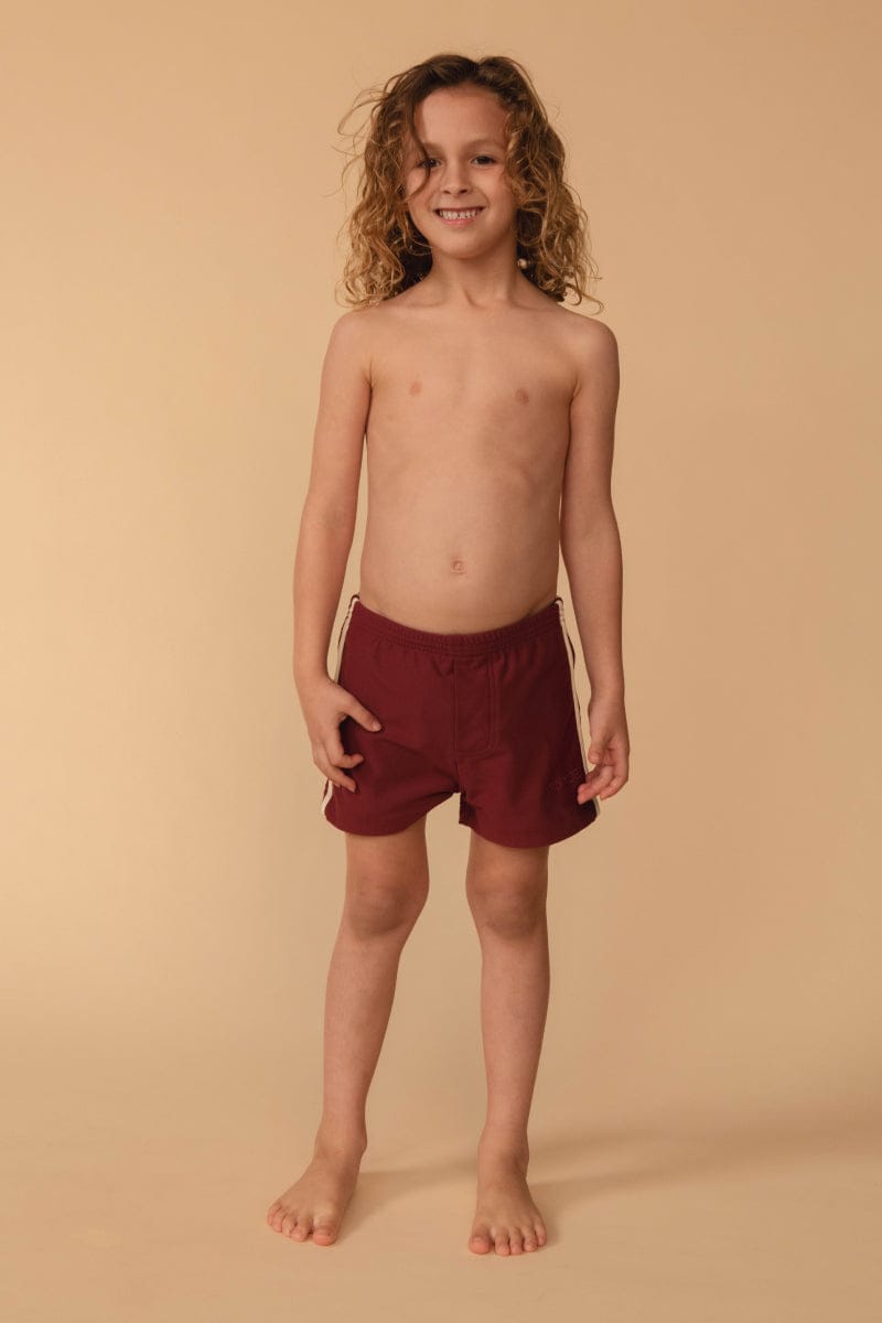 Child wearing maroon swim shorts with tan side stripes and drawstring waistband.