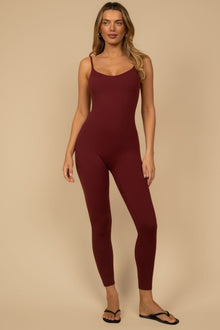 Woman wearing a maroon sleeveless jumpsuit with thin straps and full-length legs, paired with black slide sandals.