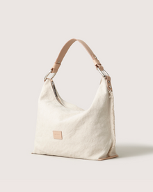 The Graf Lantz Hana Hobo is a soft, slouchy beige canvas shoulder bag with an off-white background. It has a single Vachetta leather strap attached by metal rings and features a leather patch on the front.