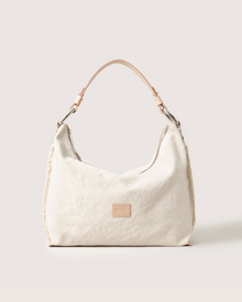 The Hana Hobo by Graf Lantz is a beige shoulder bag with a tan Vachetta leather handle, featuring a front rectangular patch. Its minimalist design and semi-slouchy structure enhance both style and functionality against a light background.
