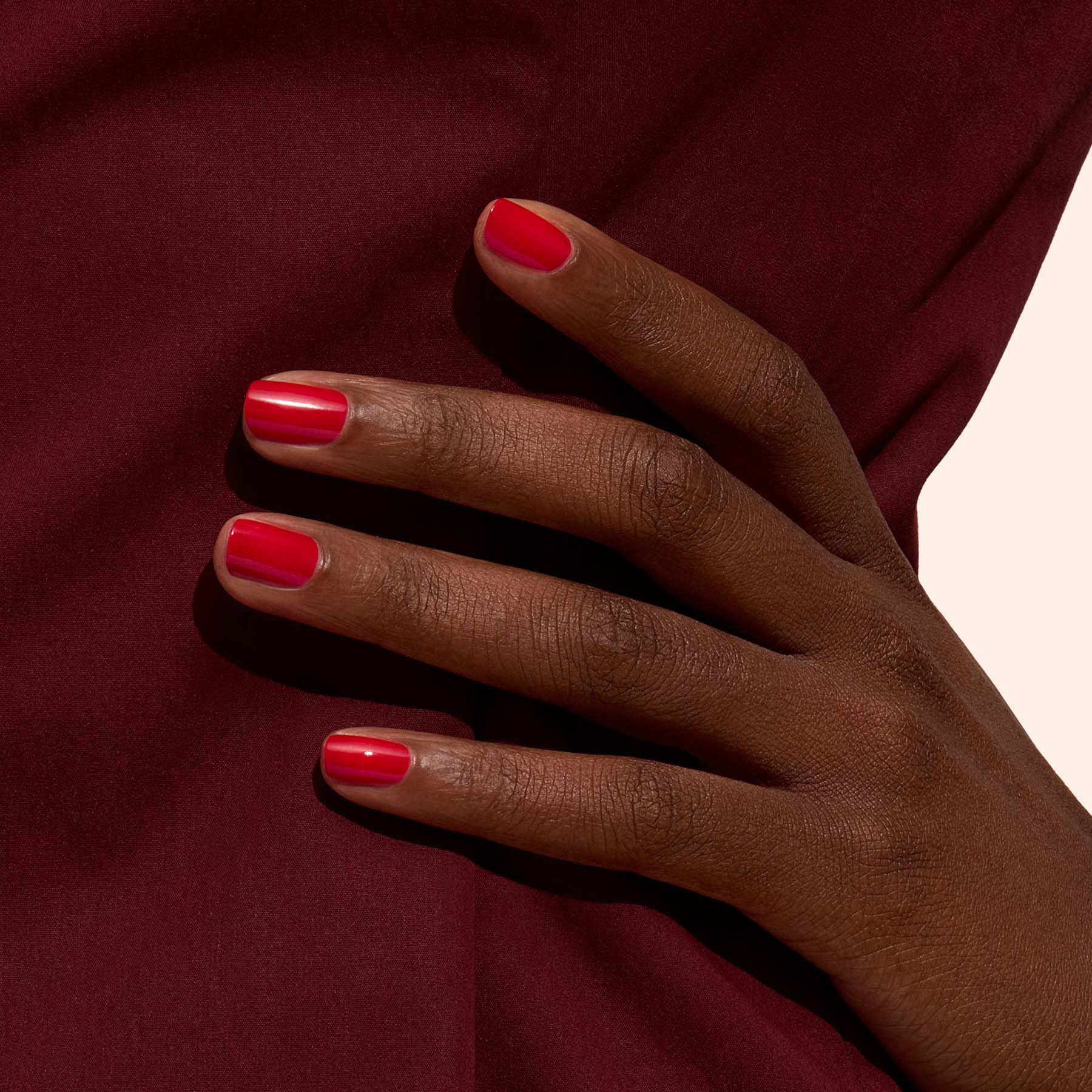 A hand with dark skin and neatly manicured nails painted in Manucurist’s Poppy Red rests against a burgundy fabric background.