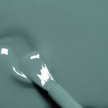 A close-up of a brush applying glossy teal nail polish on a smooth surface, with light reflections visible on the wet polish.